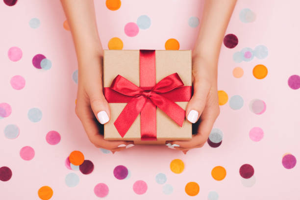 Woman hands holding present box with red bow on pastel pink background with multicolored confetti. Flat lay style.